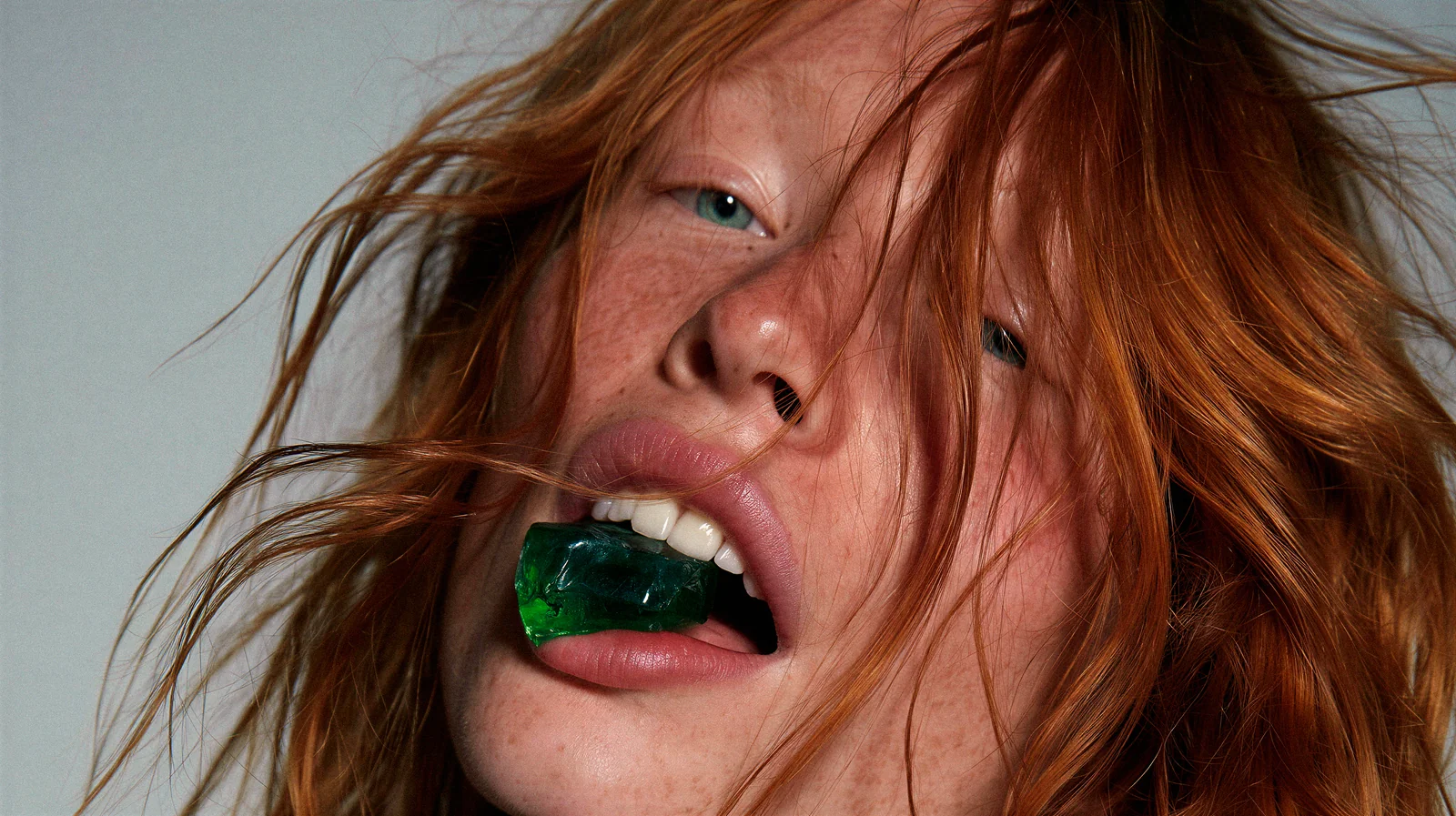 Close-up portrait of a freckled red-haired woman holding an emerald-green gem between her teeth